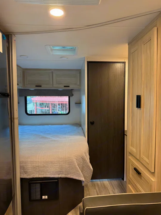 Explore this RV rental's cozy bedroom featuring a comfortable bed, light-toned cabinets, and a dark wood door, ideal for travel.. Forest River Sunseeker LE 2023