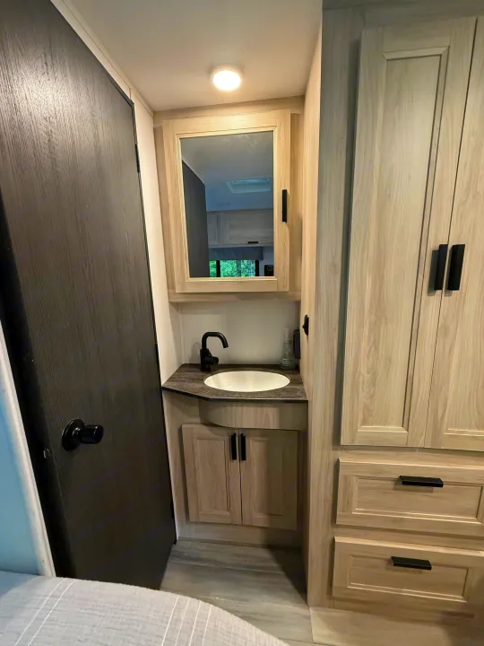 Modern RV rental vanity area featuring a sink, mirror, black faucet, and light wood cabinetry with storage. Dark door on left.. Forest River Sunseeker LE 2023