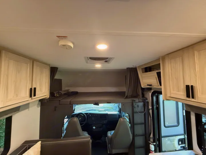 View into an rv rental's cab-over bunk and cockpit. Light wood cabinets, a vent, and a smoke detector are visible inside.. Forest River Sunseeker LE 2023