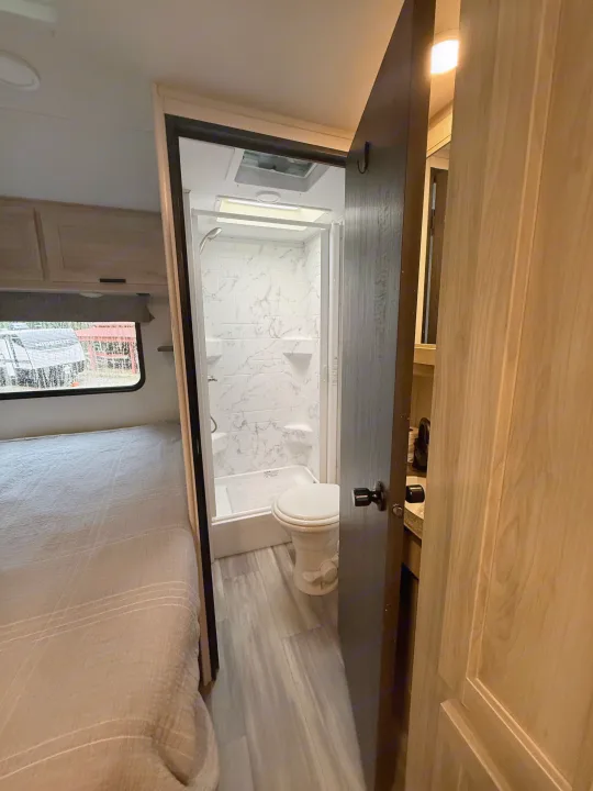 An rv rental interior featuring a compact bathroom with shower and toilet, seen from the adjacent bedroom area.. Forest River Sunseeker LE 2023