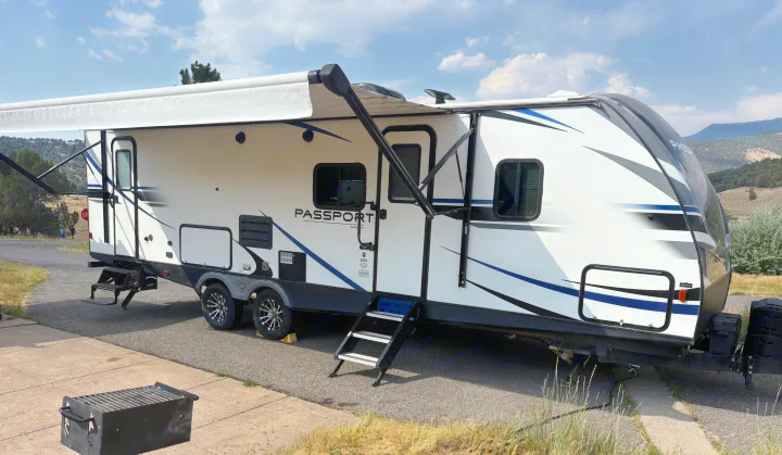 A white Passport travel trailer rv rental set up at a sunny campsite with an extended awning, steps, and grill. Mountains in the background.. Keystone RV Passport Grand Touring 2020