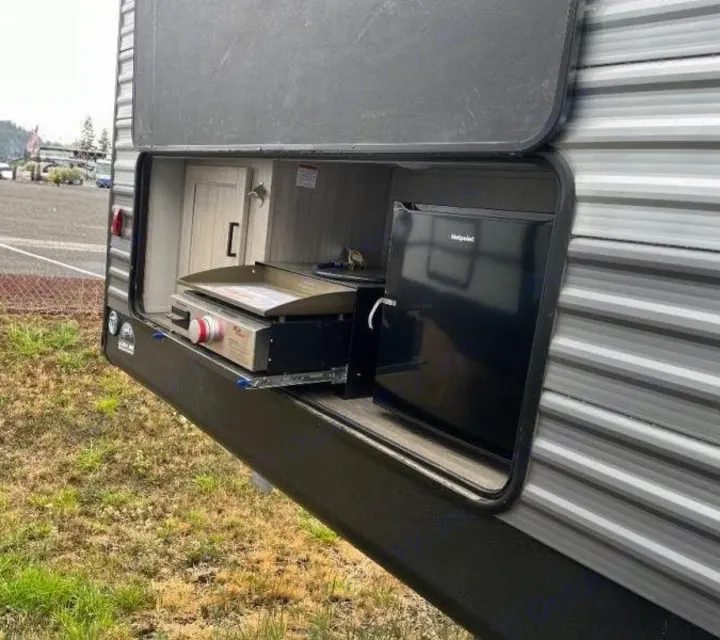 An RV rental's exterior storage compartment open, revealing a pull-out griddle and a Hotpoint mini-fridge.. Forest River Catalina Coachmen Legacy 293QBCKLE 2026