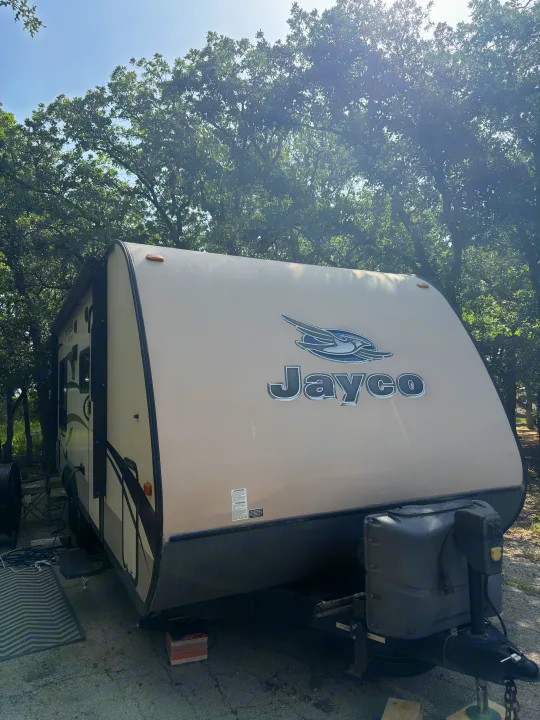 A tan Jayco travel trailer, ideal for an rv rental, is parked at a sunny campsite under lush green trees. RV is ready for adventure.. Jayco Jay Feather Ultra Lite 2015