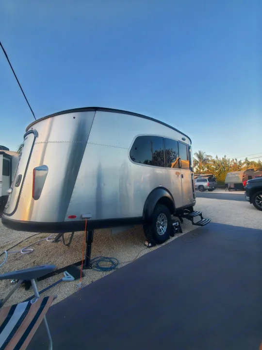 Modern silver teardrop RV rental with rugged tires parked at a sunny campsite. Utility cords and ground mat visible.. Airstream Basecamp 2022