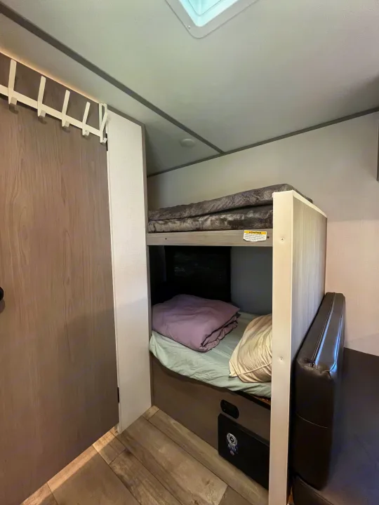 RV rental interior: stacked bunk beds with gray and purple comforters, a window, and a dark brown upholstered seat.. Coachmen 17b 2024