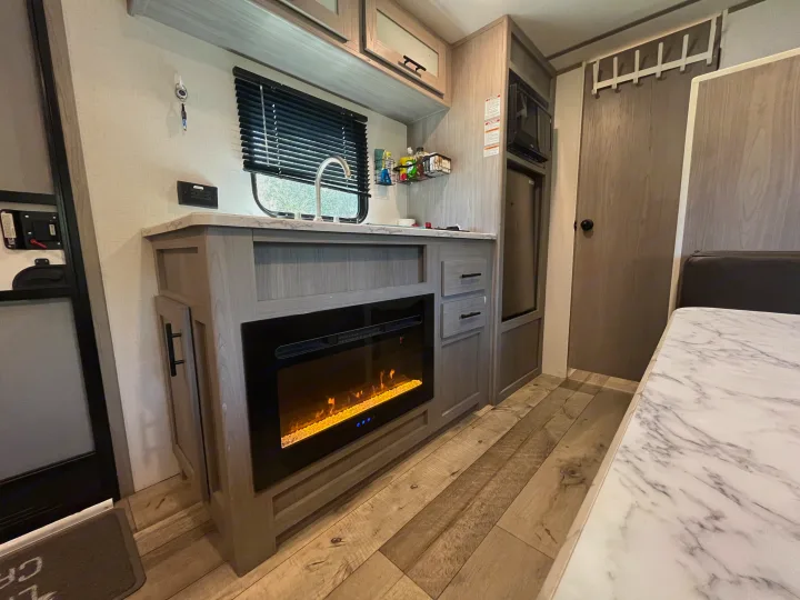 Inside a modern **rv rental**: a compact kitchen area with a sink, microwave, fridge, and cozy electric fireplace.. Coachmen 17b 2024