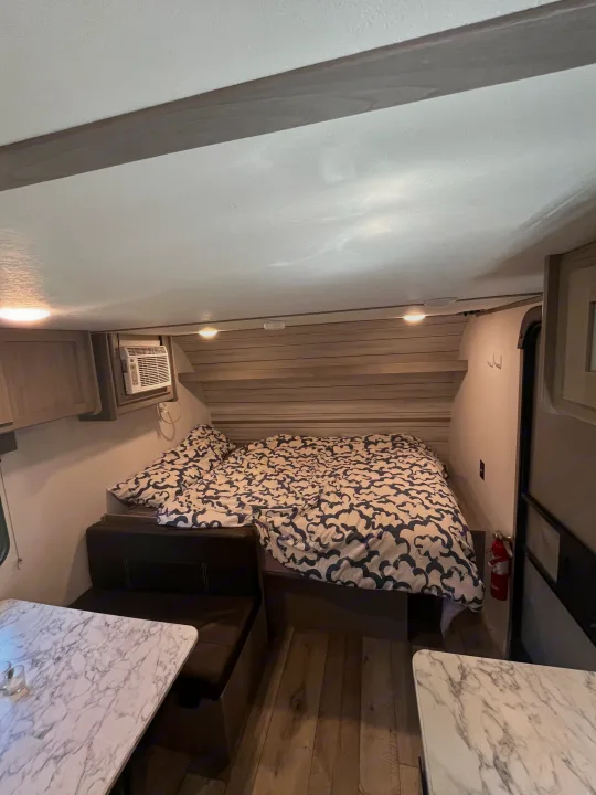 Cozy rv rental interior with a bed, patterned duvet, compact AC unit, and dinette seating with marble-look tables.. Coachmen 17b 2024