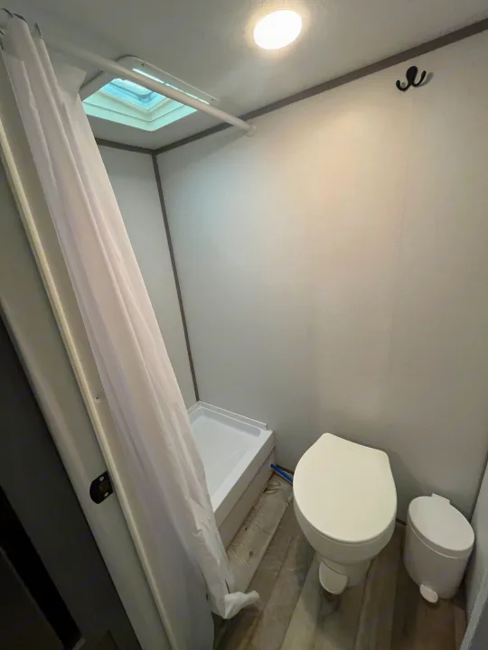 Clean RV rental bathroom with shower, toilet, and hooks. Features a skylight, overhead light, and wood-look floor.. Coachmen 17b 2024