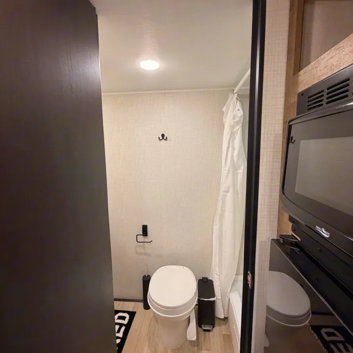 Inside an RV rental, this compact bathroom features a toilet, shower with curtain, and hooks. Microwave is visible on the right.. Keystone RV Springdale Mini 2026