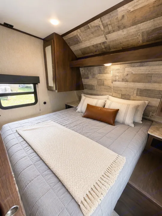 This cozy RV rental bedroom features a comfy bed with grey bedding, rustic wood paneling, and overhead storage, perfect for travel.. Dutchmen Coleman Lantern LT 2021