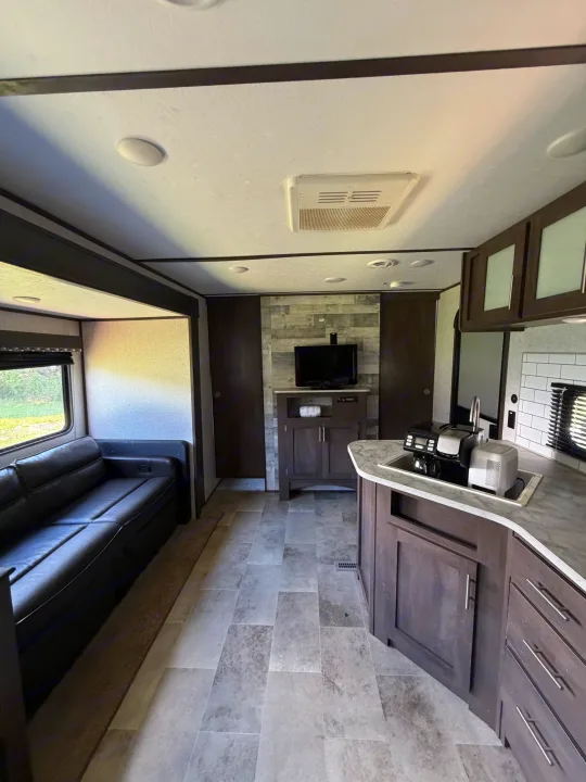 Clean rv rental interior: dark sofa, full kitchen with sink/coffee maker, TV, wood accents, and modern grey floor.. Dutchmen Coleman Lantern LT 2021