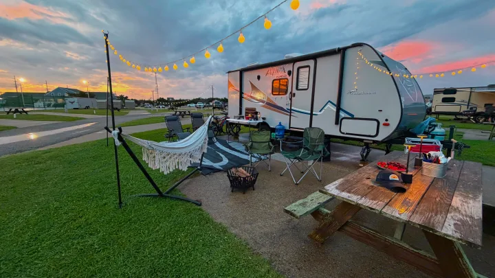 Relaxing RV rental campsite at sunset: string lights illuminate a travel trailer, hammock, picnic table, and chairs.. Coachmen Apex 2017