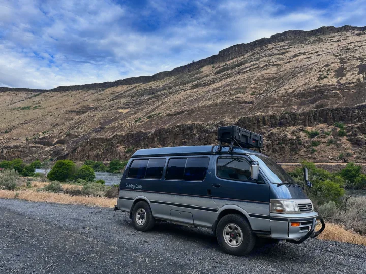 This adventure-ready blue camper van, ideal for an rv rental, is parked beside a scenic river with a huge rocky canyon.. Toyota Hiace Campervan 1994