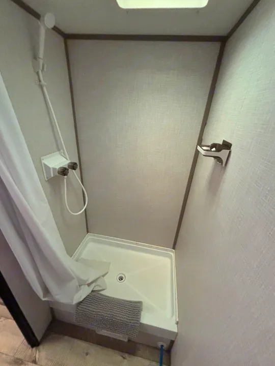 Compact RV rental shower stall with a white curtain, handheld shower, water controls, and a gray bath mat.. Dutchmen Coleman Lantern LT 2023
