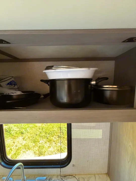 RV rental kitchen: an open cabinet with stacked pots, pans, and white colander. Below, a window shows vibrant green grass.. Dutchmen Coleman Lantern LT 2023