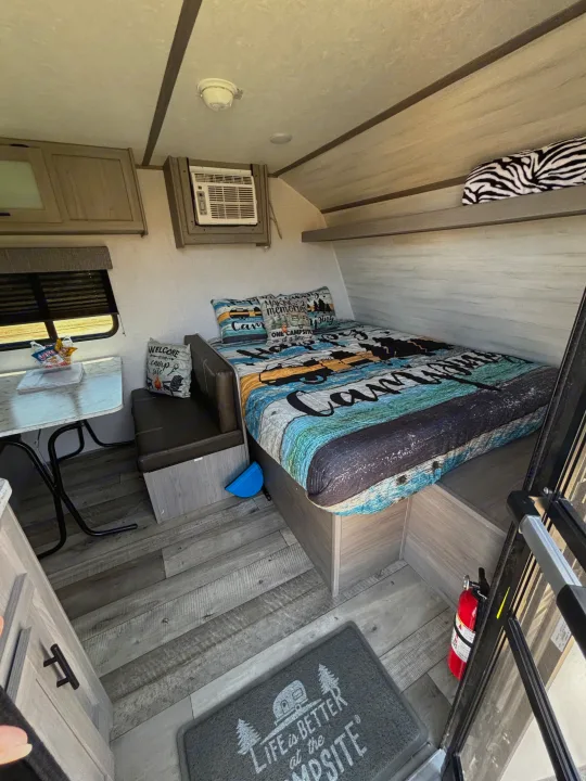 This RV rental interior features a cozy bed with camping linens, a dinette, AC unit, and a fire extinguisher for safe travels.. Dutchmen Coleman Lantern LT 2023