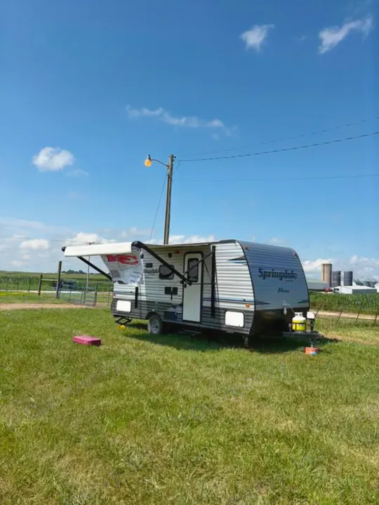Sunny day with an RV rental travel trailer on grass, awning open, a utility pole, and distant farm buildings.. Keystone RV Springdale Eco-Lite 2018