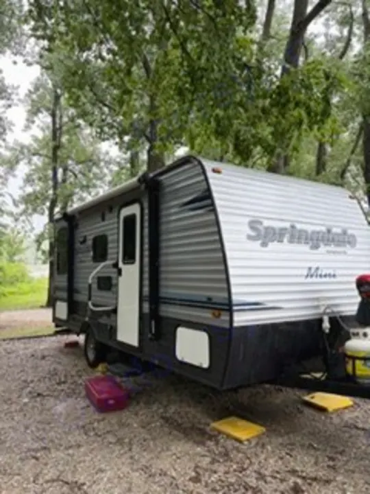 This gray and white Springdale Mini rv rental is parked on gravel under lush green trees. Ready for your camping trip!. Keystone RV Springdale Eco-Lite 2018