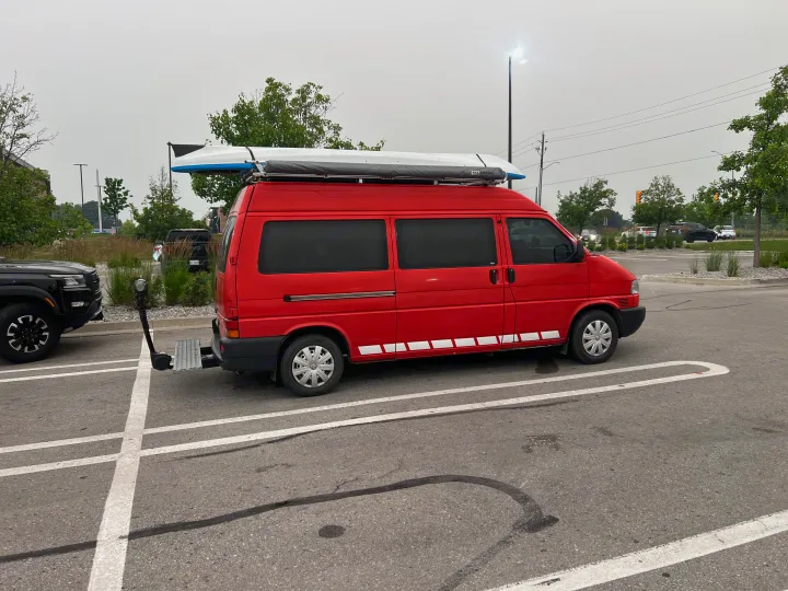 Bright red custom camper van with rooftop kayak and rear access platform parked in a lot. Ideal RV rental for travel.. Volkswagen Multivan 2003