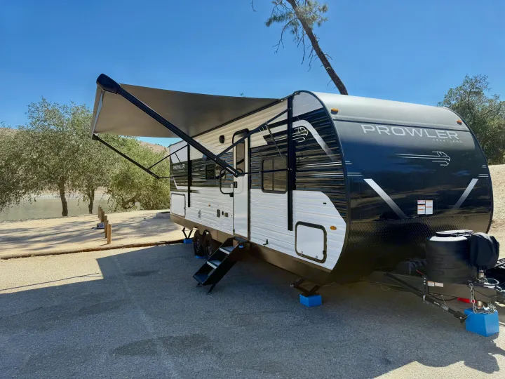 This Prowler Heartland travel trailer rv rental is set up at a scenic lakefront campground with its awning extended.. Heartland Powler 2026