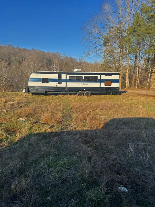 White and blue RV rental travel trailer parked in a sunny, dry field with a forest and clear blue sky.. Forest River Cherokee Grey Wolf 2026