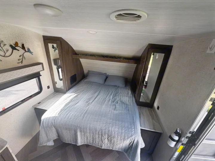 RV rental bedroom with a gray quilted bed, mirrored wardrobes, window, overhead shelf, ceiling lighting, and air vent.. Forest River Wildwood X-Lite 2021