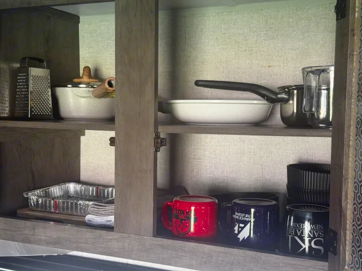 RV rental kitchen cabinet showing organized pots, a grater, and branded mugs from Taos, Arapahoe Basin, and Santa Fe.. Forest River Wildwood X-Lite 2021