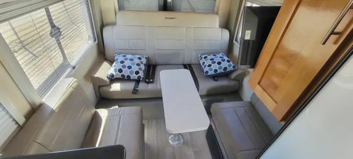 This bright rv rental shows a dinette area with tan benches, a white table, and blue geometric pillows by the window.. Coachmen Crossfit 2019