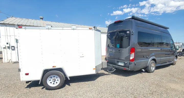 Grey Ford Transit **rv rental** towing a white utility trailer on a gravel lot under a clear blue sky.. Coachmen Crossfit 2019