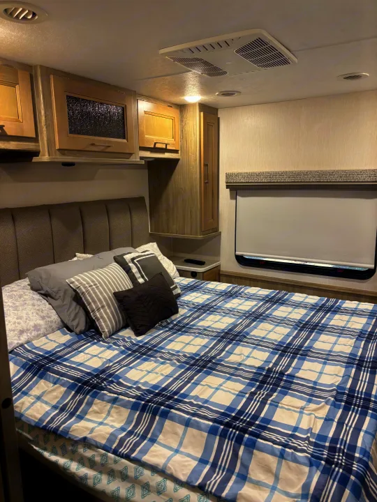 Inside a comfy RV rental bedroom: plush bed, plaid comforter, storage cabinets, and window with shade.. Coachmen Mirada 32ss 2020