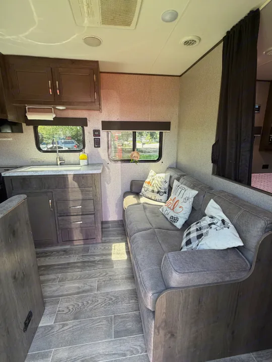 Bright RV rental living space featuring a cozy gray sofa with fall pillows, a kitchen area, and wood-look flooring.. Jayco Jay Flight SLX 2021
