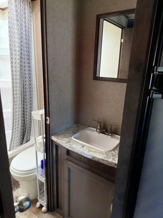 Compact RV rental bathroom featuring a shower with a patterned curtain, toilet, sink vanity, and mirror, showing a functional RV interior.. Jayco Jay Flight SLX 2021