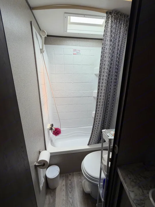 Compact rv rental bathroom: shower/tub with grey patterned curtain, toilet, and toilet paper holder. A bright skylight is overhead.. Jayco Jay Flight SLX 2021