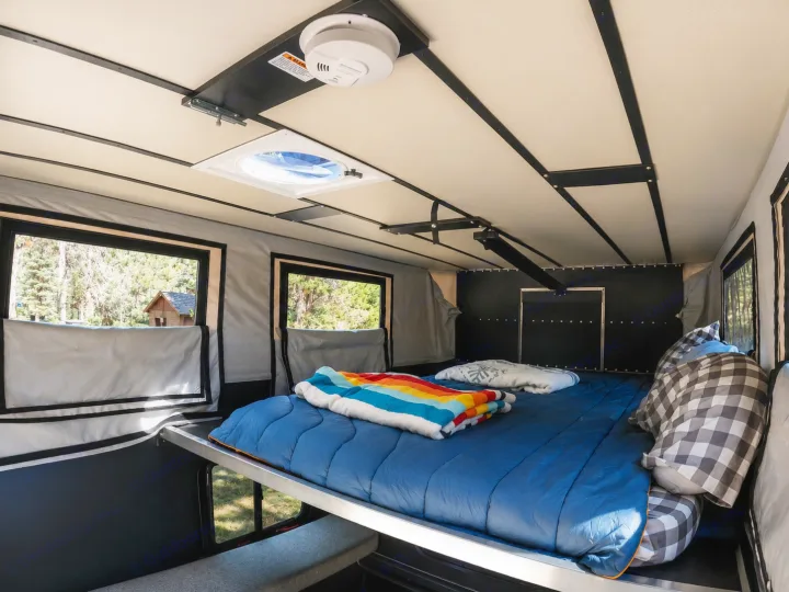 Bright rv rental interior showing a comfortable bed with a colorful blanket, windows with forest views, and a ceiling vent.. Four Wheel Campers Shell Models 2020