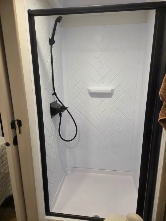 Modern, compact white shower with black fixtures inside an RV rental, featuring a herringbone pattern wall and a small shelf.. Alliance RV Delta 2026