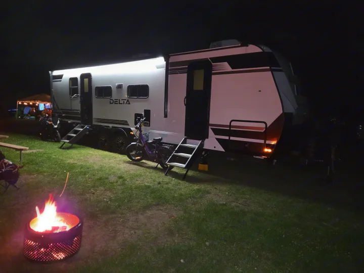 Nighttime rv rental scene with a brightly lit RV, campfire, bikes, and a social gathering, perfect for camping trips.. Alliance RV Delta 2026