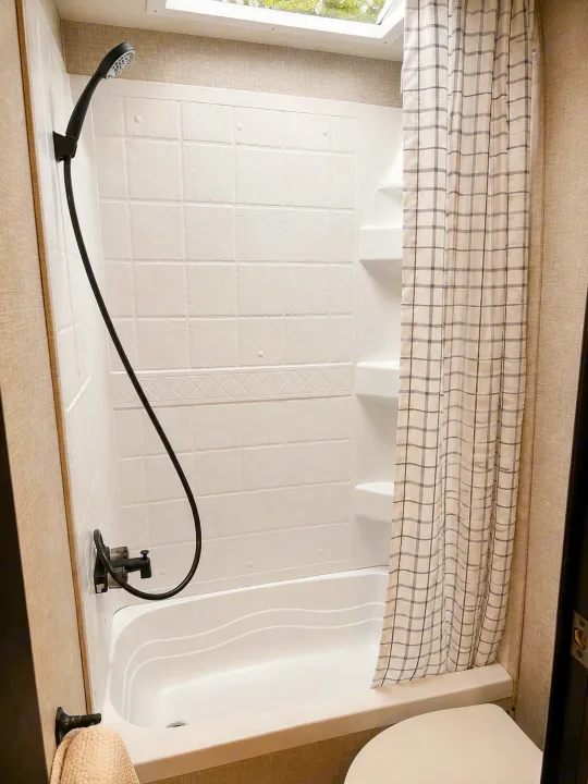 Compact RV rental bathroom with white shower/tub, black fixtures, plaid curtain, and skylight. Ideal for travel.. Jayco Jay Flight SLX 2025