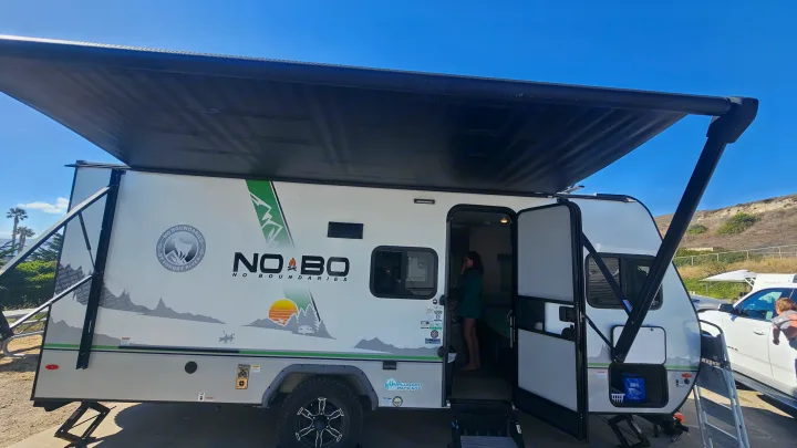 This "NO BOUNDARIES" travel trailer RV rental, with its black awning extended, is parked outdoors. A person stands inside the open doorway.. No Boundaries 19 2022