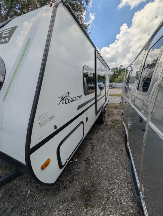 A white Coachmen travel trailer and a shiny silver RV side-by-side in a gravel lot under a cloudy sky, ready for rv rental.. Coachmen Other 2024