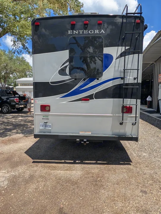 Rear view of an Entegra RV rental, featuring a ladder, brake lights, and a Quebec license plate. A person works on a pickup.. Entegra Coach Vision XL 2022