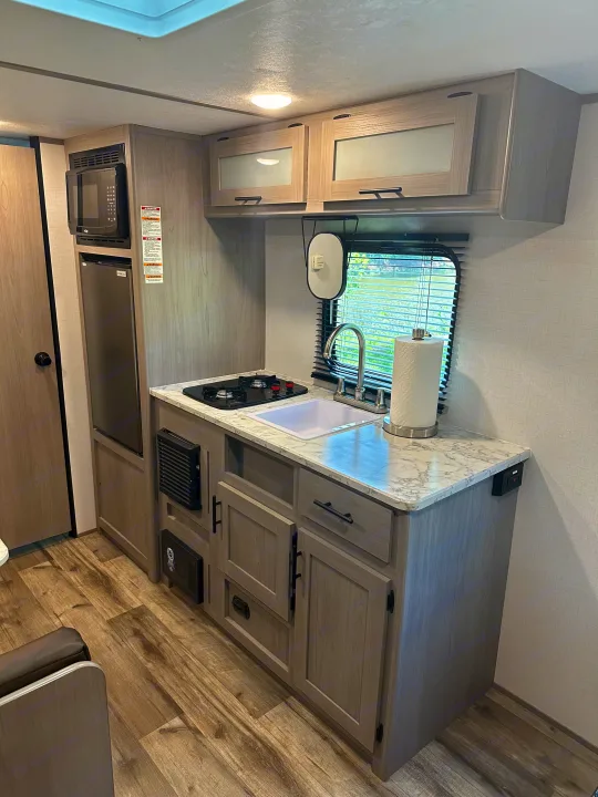 Modern RV rental kitchen with microwave, fridge, stove, sink, marble counters, light wood cabinets, and wood floors.. Keystone RV Aspen trail 2024