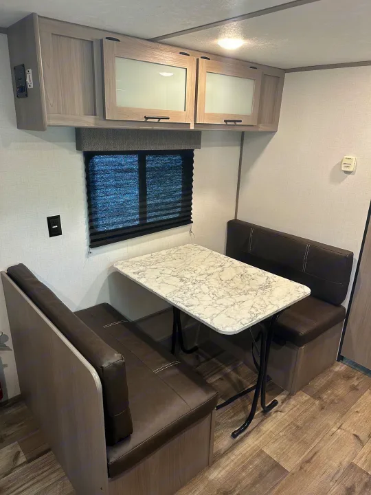 Cozy rv rental dinette with a marble table, dark brown bench seating, overhead cabinets, and a window with blinds.. Keystone RV Aspen trail 2024
