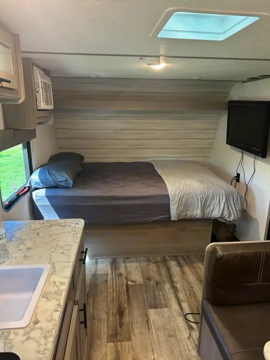 Modern rv rental interior showing a cozy bedroom with bed, AC, TV, and a compact kitchen counter with sink.. Keystone RV Aspen trail 2024