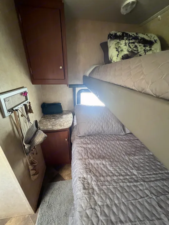 Cozy RV rental interior with bunk beds, quilted gray bedding, wooden cabinetry, and a bright window. Wall organizer visible.. Forest River Wildwood X-Lite FS 2015
