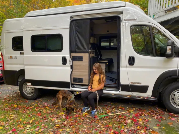 Girl and dog by a white rv rental, enjoying autumn leaves. Perfect for family travel in a camper van.. Winnebago Solis 2023