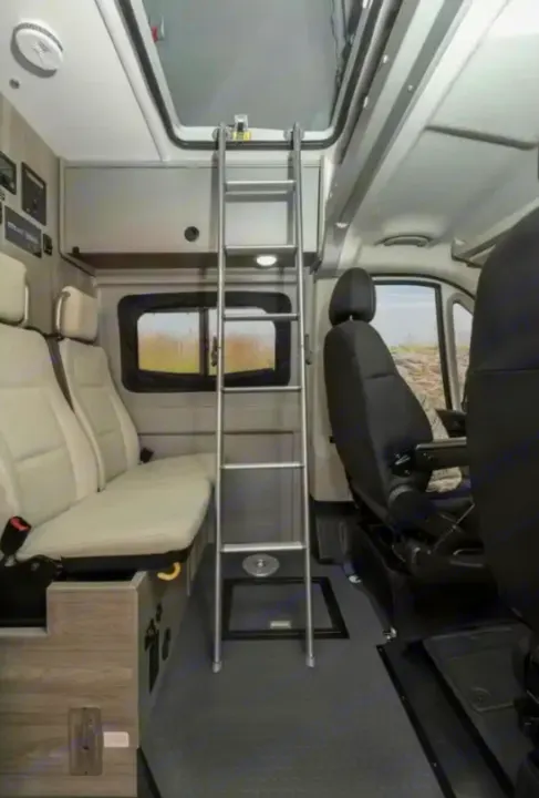 This RV rental interior shows a ladder to an overhead bed, passenger seats, and the front cockpit area of a camper van.. Winnebago Solis 2023