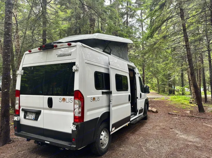 This white Winnebago Solis rv rental, with its pop-top open, is perfectly parked on a forest path, ready for adventure.. Winnebago Solis 2023