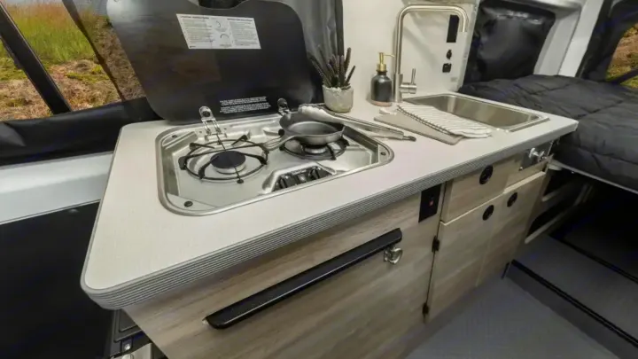 Experience comfort in this RV rental's compact kitchen with a two-burner stove, sink, and integrated storage, adjacent to a bed.. Winnebago Solis 2023