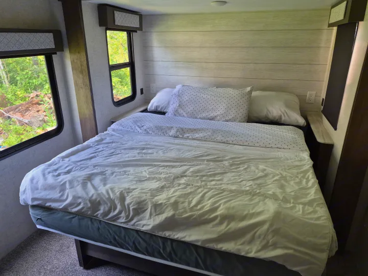 Cozy RV rental bedroom with a comfy bed, white and dotted bedding, light wood paneling, and windows showing green trees.. Forest River Wildwood 2021