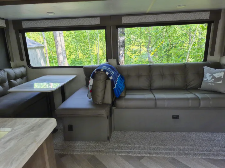 This RV rental features a comfortable dinette and sofa, with large windows overlooking a lush green view. Perfect for camping.. Forest River Wildwood 2021